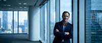 Confident middle-aged Caucasian businessman with crossed arms standing in modern office with city skyline view through floor-to-ceiling windows.