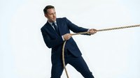 Determined businessman in navy suit pulling rope in tug of war competition showing leadership strength and perseverance in corporate challenge.