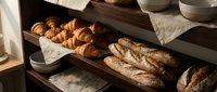 Fresh baked artisan bread and croissants displayed on wooden shelves in warm bakery lighting for commercial food photography and culinary marketing.