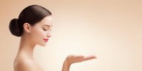 Profile view of Asian woman with sleek bun hairstyle and red lips presenting with open palm against soft beige background for beauty product advertisement.
