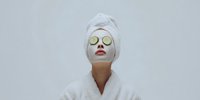 Woman in white bathrobe and towel with cucumber slices on eyes enjoying spa treatment against minimalist background. Perfect for wellness advertising.