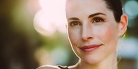 Portrait of mature woman with natural makeup smiling at sunset, warm golden light creating soft bokeh background.