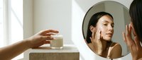 Young woman with olive skin tone examining her face in round mirror, touching cheek while applying skincare product. Suitable for beauty, cosmetics, wellness and dermatology marketing materials.