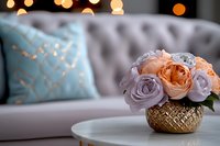 Fresh peach garden roses and lavender roses arranged in textured gold vase on marble table, with gray tufted sofa and blue pillow in soft bokeh background.