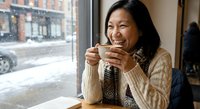 Happy Asian woman enjoying hot beverage in cozy cafe during winter, smiling while holding warm drink cup near window with snowy street view outside.