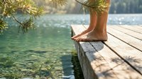 Woman relaxing barefoot on wooden dock by crystal clear mountain lake surrounded by pine trees during peaceful summer vacation retreat.
