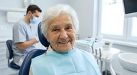 Happy elderly Caucasian woman patient smiling in dental chair during checkup with masked dentist in modern clinic office for oral health care treatment.