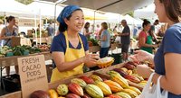 Asian female vendor in yellow apron selling fresh cacao pods at outdoor farmers market, engaging with customer in community commerce setting.