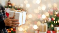 Senior man holding wrapped gift box with gold ribbon during festive holiday celebration with decorated tree and warm bokeh lights in background.