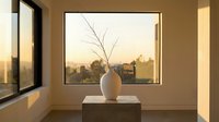 Minimalist interior with white ceramic vase and bare branches on wooden table against window overlooking urban cityscape at golden hour sunset.