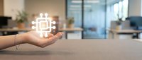 Hand holding glowing microchip processor icon in modern office workspace representing artificial intelligence and technology innovation.