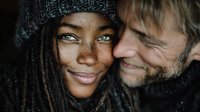 Young African American woman with dreadlocks and knit hat embracing smiling Caucasian man with beard in intimate winter portrait for relationship concepts.