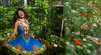 Young woman with dark curly hair and medium brown skin tone wearing traditional African print blue dress and red patterned scarf, laughing joyfully in lush garden surrounded by blooming flowers and green foliage.