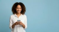 Happy young African American woman with curly hair using smartphone on blue background for social media marketing and communication concepts.