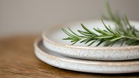 Fresh rosemary sprig on rustic ceramic plates with textured edges against wooden surface, soft focus background creates minimalist dining atmosphere.