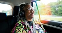 Happy African American woman with braids wearing headphones enjoying music while traveling as passenger in car with sunlight streaming through window.