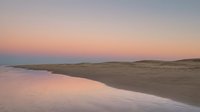 Serene beach landscape at sunset with soft pink and purple sky reflecting in calm water along sandy shoreline creating peaceful coastal scenery for relaxation themes.