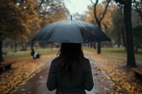 Woman with black umbrella walking alone in autumn park on rainy day, fallen orange leaves on wet path create moody atmospheric scene. Selective focus view from behind.