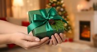 Green wrapped gift box with emerald ribbon and pine cone decoration held in hands during festive holiday season with warm fireplace background.