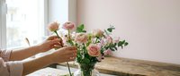 Woman arranging fresh pink roses and greenery in glass vase on wooden table by window. Hands placing flowers in bright natural light interior setting.