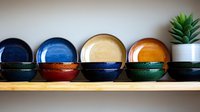 Colorful ceramic bowls and plates arranged on wooden shelf with small potted succulent plant, showcasing artisanal pottery collection in blue, orange and green glazes.