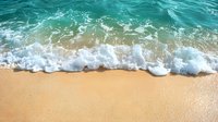 Turquoise ocean wave washing onto golden sandy beach with white foam, creating natural pattern where water meets shore. Aerial view of tropical coastline.
