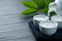 Clear liquid pouring from glass bottle into white ceramic bowls on dark slate with green leaves, spa wellness aromatherapy skincare treatment concept.