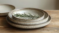 Handmade ceramic plates with speckled glaze and fresh rosemary sprig on wooden table for rustic kitchen decor and natural dining presentation.