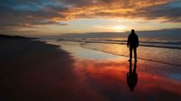 Silhouetted surfer holding board standing on wet beach sand at golden sunset with dramatic sky and ocean waves, suitable for travel and lifestyle content.