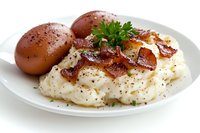 Creamy mashed potatoes topped with crispy bacon bits and fresh parsley, served with seasoned brown eggs on white plate against light background.