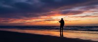 Silhouette of fisherman casting line at ocean beach during dramatic sunset with colorful sky reflecting on wet sand and gentle waves rolling ashore.