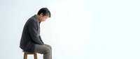 Young Asian businessman in dark suit sitting on wooden stool looking down in contemplative pose against white background for business concepts.
