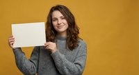 Young woman with curly brown hair holding blank white paper card against yellow background, smiling at camera wearing gray knit sweater for mockup design.