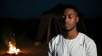 Young African American male, casual worker or laborer, wearing light-colored shirt, contemplative expression, sitting near campfire at night. Suitable for themes of solitude, reflection, outdoor work, or evening leisure activities.