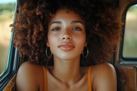 Young African American woman with natural curly afro hair and hoop earrings smiling confidently in car backseat during golden hour lighting, wearing orange tank top.