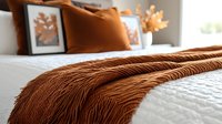 Cozy bedroom interior with rust colored textured throw blanket on white bedding, autumn home decor with orange pillows and leaf artwork creating warm atmosphere.