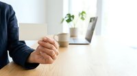 Caucasian male professional in dark blazer with clasped hands on wooden desk, laptop and potted plant in background, office workspace setting.