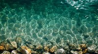 Crystal clear turquoise water with sunlight reflections creating ripple patterns on surface, rocky seabed visible through transparent Mediterranean waters.
