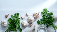 Fresh garlic bulbs, cloves, parsley, and sea salt arranged on a white wooden surface, flat lay composition for culinary and food blog use.