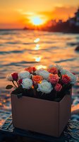 Fresh roses in brown box against dramatic ocean sunset background, waves reflecting golden light, romantic seaside setting for Valentine's Day or anniversary celebration.