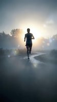 Male runner jogging on misty road at sunrise with dramatic sunlight and fog creating atmospheric morning workout scene for fitness motivation content.