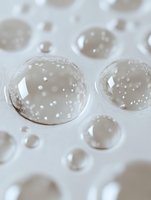 Clear water droplets on white surface create abstract macro photography pattern with light refraction and bokeh effect showing surface tension properties.