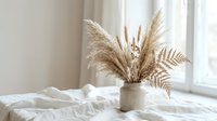 Dried pampas grass and fern leaves in ceramic vase on white bedding near window creating minimalist boho bedroom decor for interior design projects.
