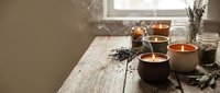 Lit candles in ceramic holders arranged on wooden table with dried lavender, pampas grass, and glass vases near window for home decor and wellness styling.