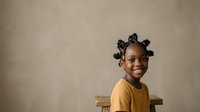 Young Black girl with braided hair buns wearing yellow shirt, sitting on wooden chair against neutral beige background. Studio portrait for children's content.