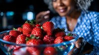 Fresh ripe strawberries in blue glass bowl with African American woman smiling in background, selective focus on vibrant red berries for food photography.