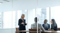 Professional businesswoman presenting to corporate team in modern glass conference room with city skyline view during executive meeting.