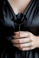 Female hands with black manicure holding dark anemone flower against black fabric background, creating dramatic gothic aesthetic mood for fashion and beauty concepts.