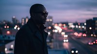Young African male professional wearing sunglasses and dark jacket standing against blurred city lights at dusk, contemplative urban portrait for business and lifestyle concepts.
