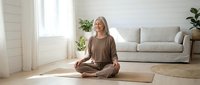 Senior woman with gray hair, Caucasian ethnicity, practicing yoga in lotus position on mat in bright living room with sofa and plants. Wellness and mindfulness concept.